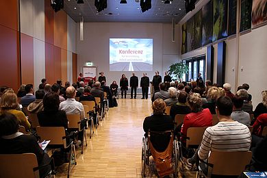 Die Perspektive richtet sich von hinten auf die Bühne. Man sieht viele Gäste von hinten, vorn singt das A-Capella-Ensemble Thonkunst.