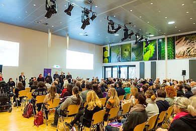 Viele Menschen sitzen in einem Raum und sind zur Bühne gerichtet